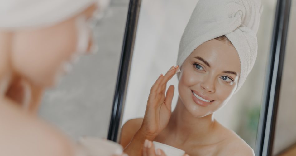 Cute woman applies beauty cream on face, smiles pleasantly, looks in mirror, poses in bathroom